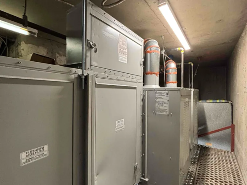 Large grey air handling units lined up along a narrow concrete mechanical room, with round flue pipes rising to the ceiling above a metal grate walkway