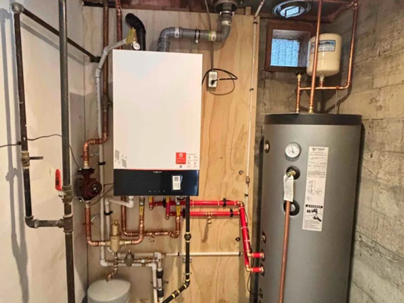 A wall-mounted boiler on a plywood backer with copper and red pipework below it, beside a tall grey storage tank in a basement
