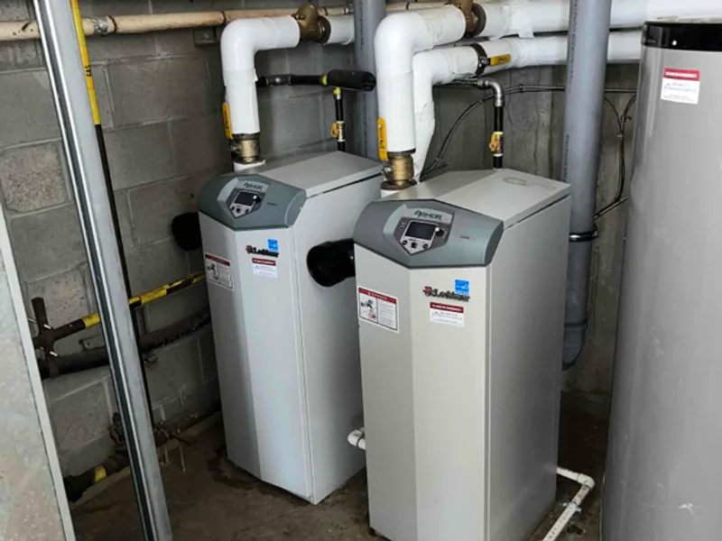 Two floor-standing boilers side by side in a concrete block mechanical room, with vent piping and copper lines running overhead