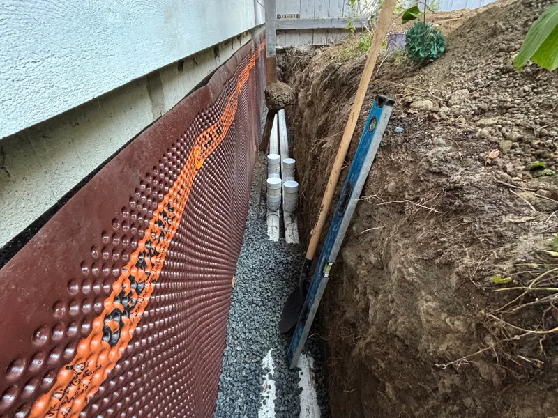 A trench opened along a house foundation, the wall covered in dimpled drainage membrane, with perforated drain pipe bedded in gravel at the bottom