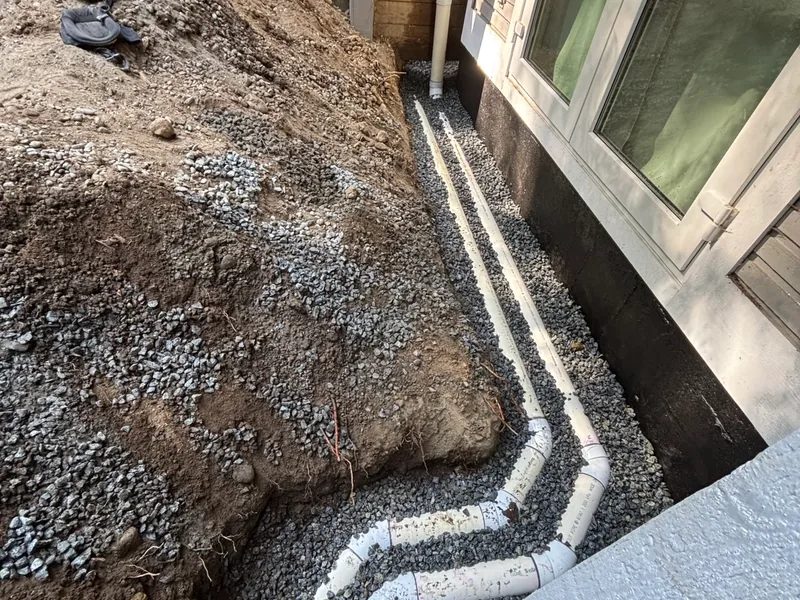 Two runs of white perforated drain pipe bedded in gravel in an open trench along a house foundation, with excavated soil piled on the far side